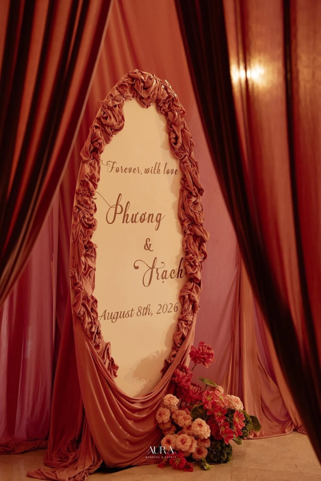 Bảng chào khung oval viền lụa hồng xếp nhún, dòng chữ Forever, with love và bó hoa hồng dưới chân, nền rèm hồng – đỏ rượu