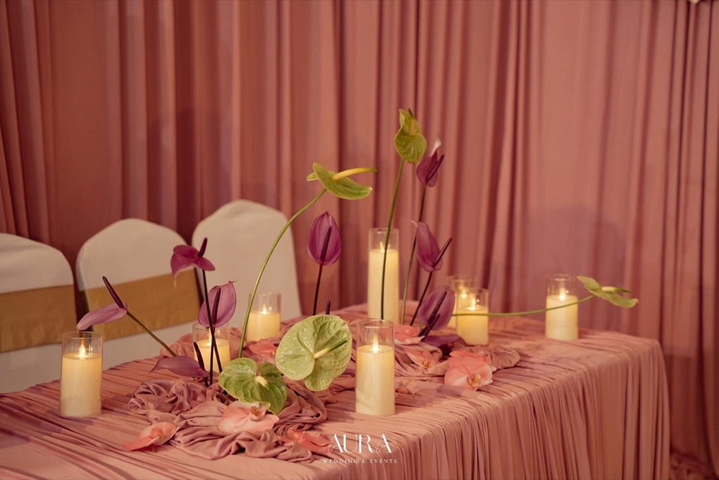 Bàn tiệc phủ lụa hồng xếp nếp với hồng môn tím – xanh vươn cao và nến trụ trong ly thủy tinh