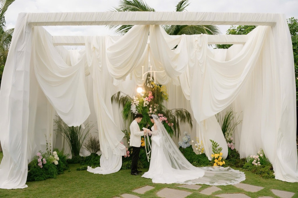Khung rạp phủ vải trắng buông rủ trên bãi cỏ, cụm hoa cỏ trung tâm dưới monogram HT, cô dâu chú rể đọc lời hứa