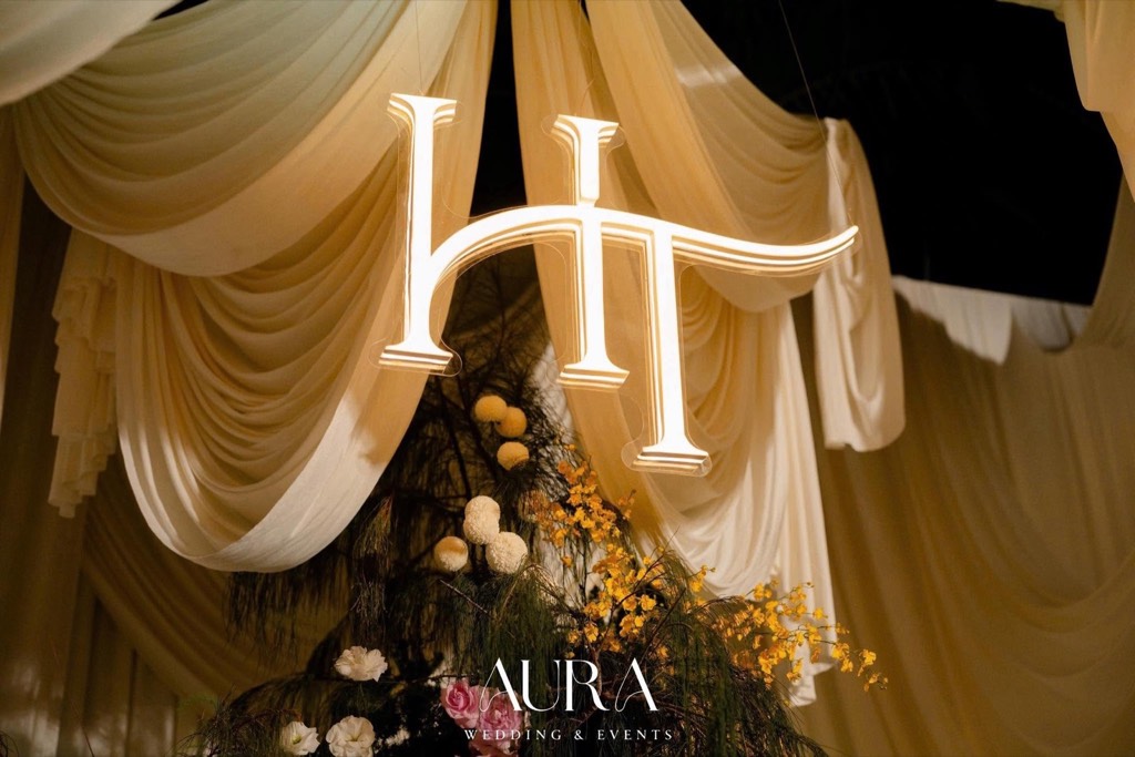 Monogram HT phát sáng giữa các lớp vải ngà xếp nếp, điểm lan vũ nữ vàng và cúc ping pong trắng