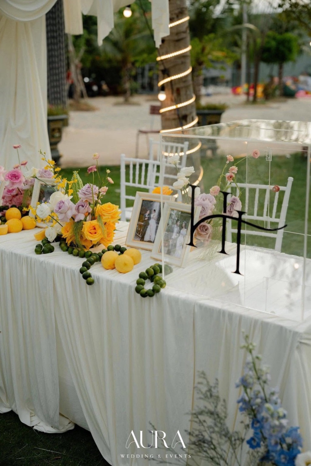 Bàn ảnh phủ vải trắng xếp nếp với hồng vàng, hồng tím, chanh vàng, chuỗi chanh xanh và hộp mica monogram HT