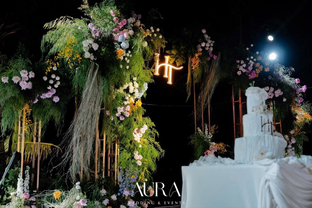 Cổng hoa khung gỗ về đêm với hồng, cẩm tú cầu, cỏ lau rủ, monogram HT phát sáng và bánh cưới trắng