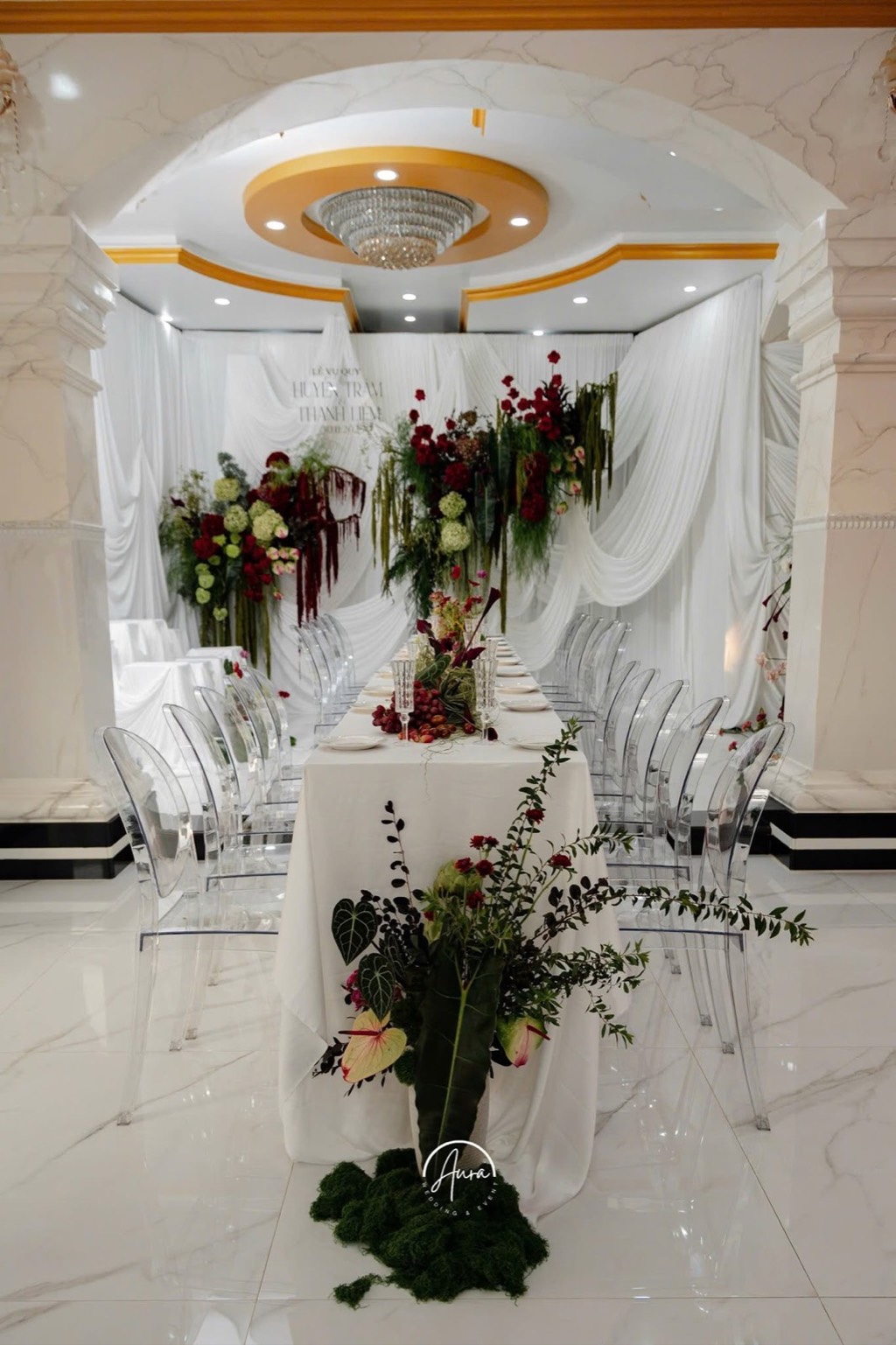 Sảnh tiệc dưới đèn chùm pha lê: bàn dài phủ khăn trắng, cụm hoa đỏ và xanh trên nền vải trắng rủ, chân bàn phủ rêu