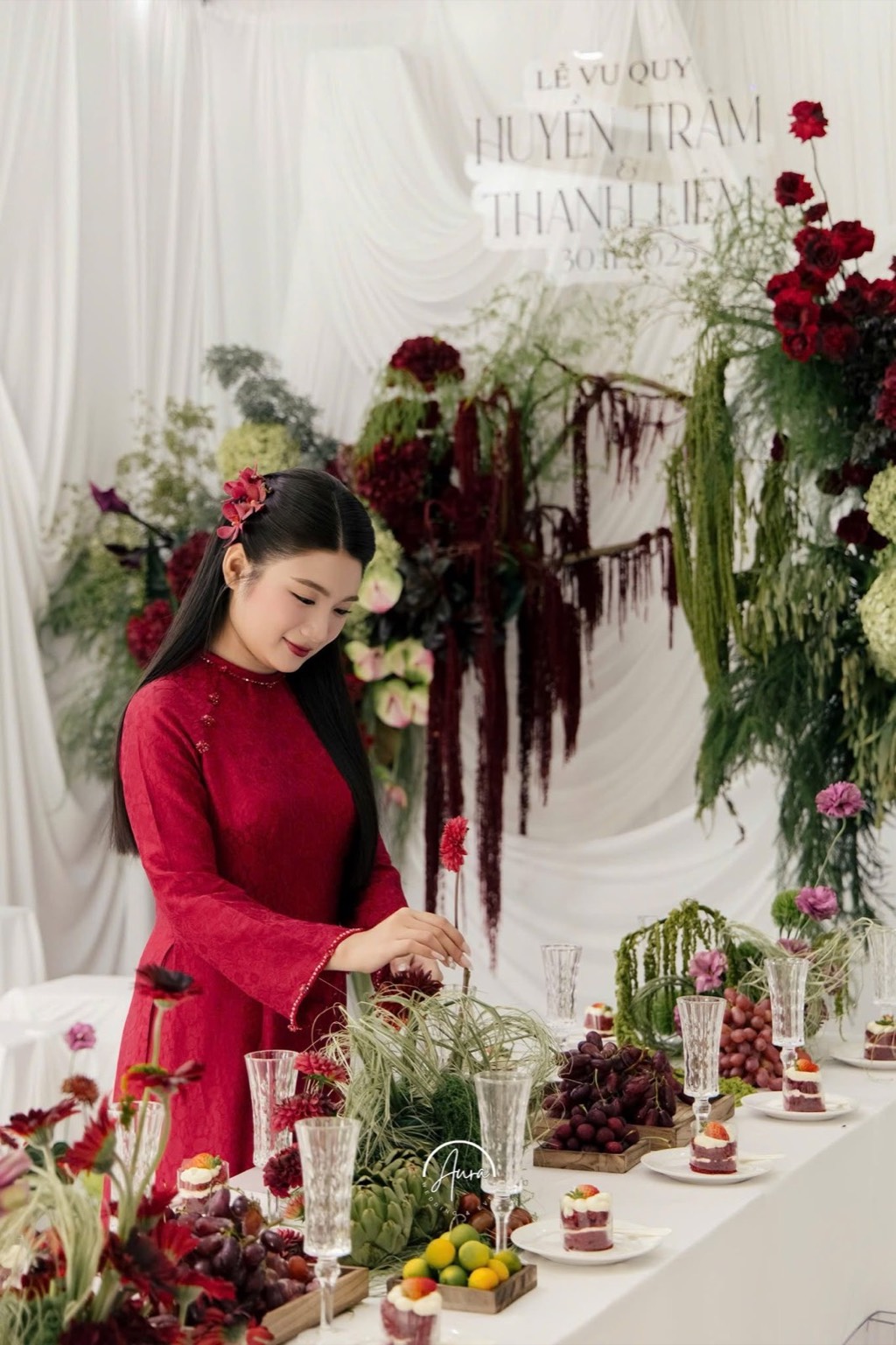 Cô dâu áo dài đỏ chỉnh bàn tiệc bày trái cây và ly pha lê, cụm hoa đỏ và olive phía sau