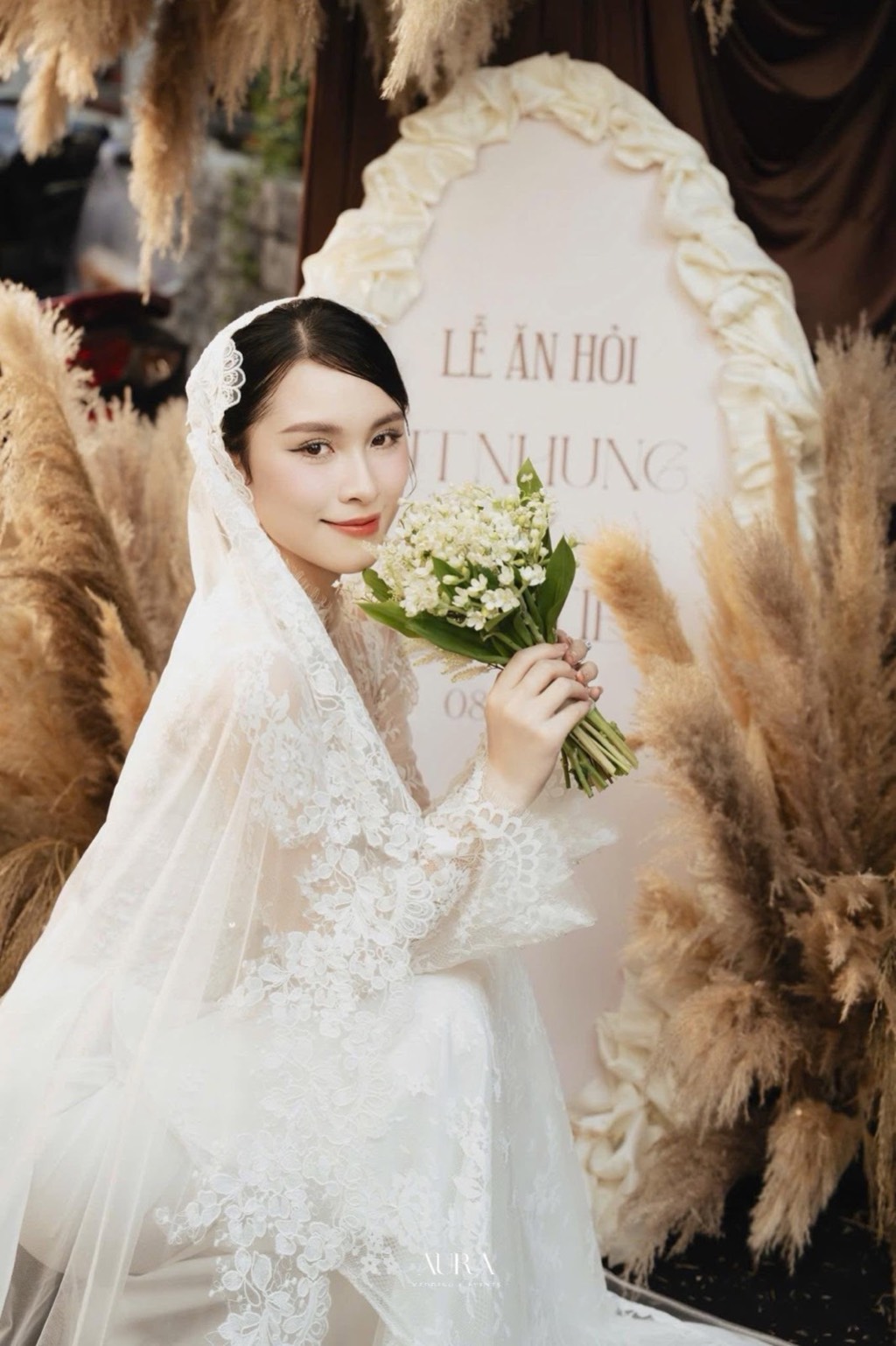 Bảng tên lễ ăn hỏi viền lụa kem xếp nếp giữa những cụm cỏ pampas, cô dâu cầm bó hoa trắng