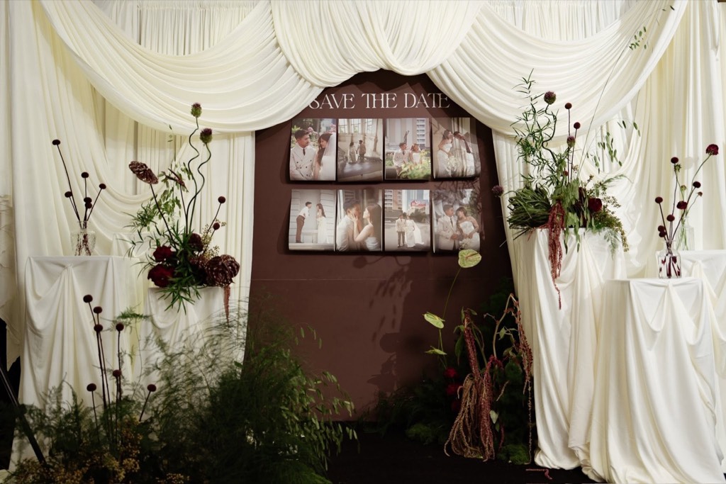 Góc Save the Date nền nâu khung lụa trắng rủ, bục phủ vải với bình hoa đỏ và cỏ xanh