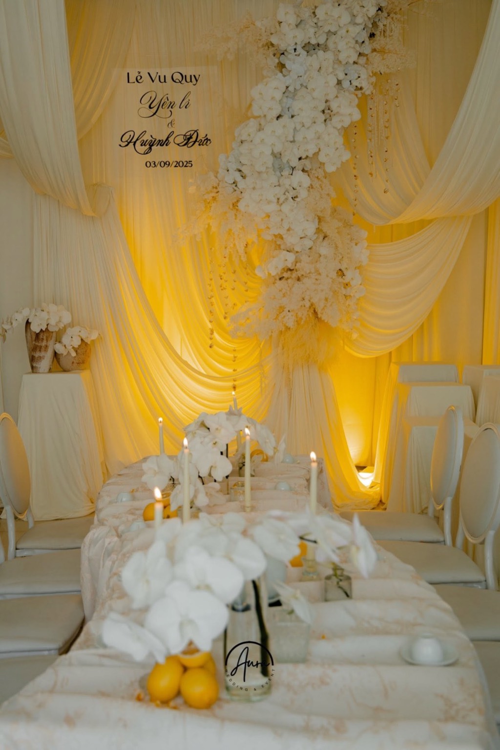 Bàn gia tiên với nến cây trắng, lan hồ điệp và chanh vàng hướng về backdrop vải ngà rực ánh vàng ấm