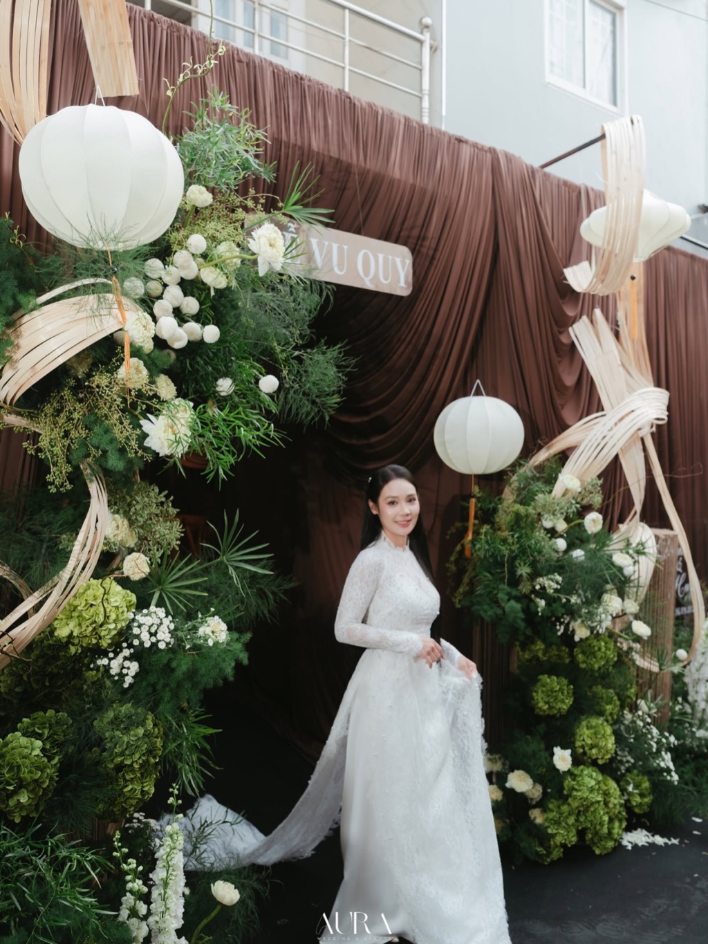 Cổng vu quy trước nhà: phông vải nâu, hai trụ hoa trắng và cẩm tú cầu xanh, vòng mây uốn và đèn lồng trắng, cô dâu áo dài trắng