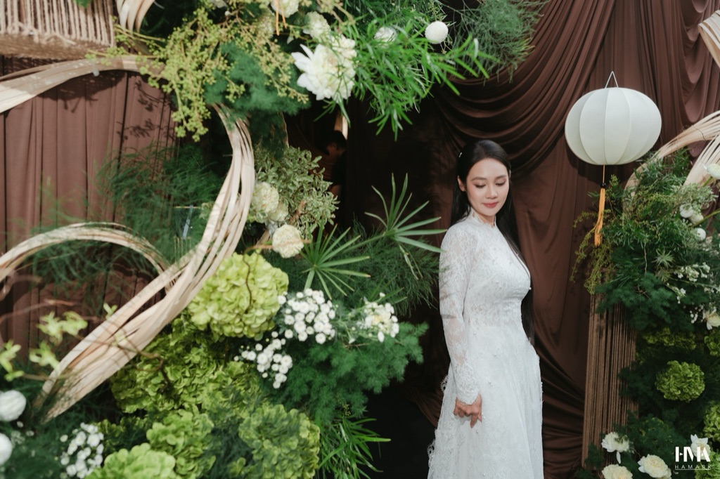 Cô dâu áo dài trắng giữa hai khối hoa xanh trắng, vòng mây uốn và đèn lồng trắng trước rèm nâu