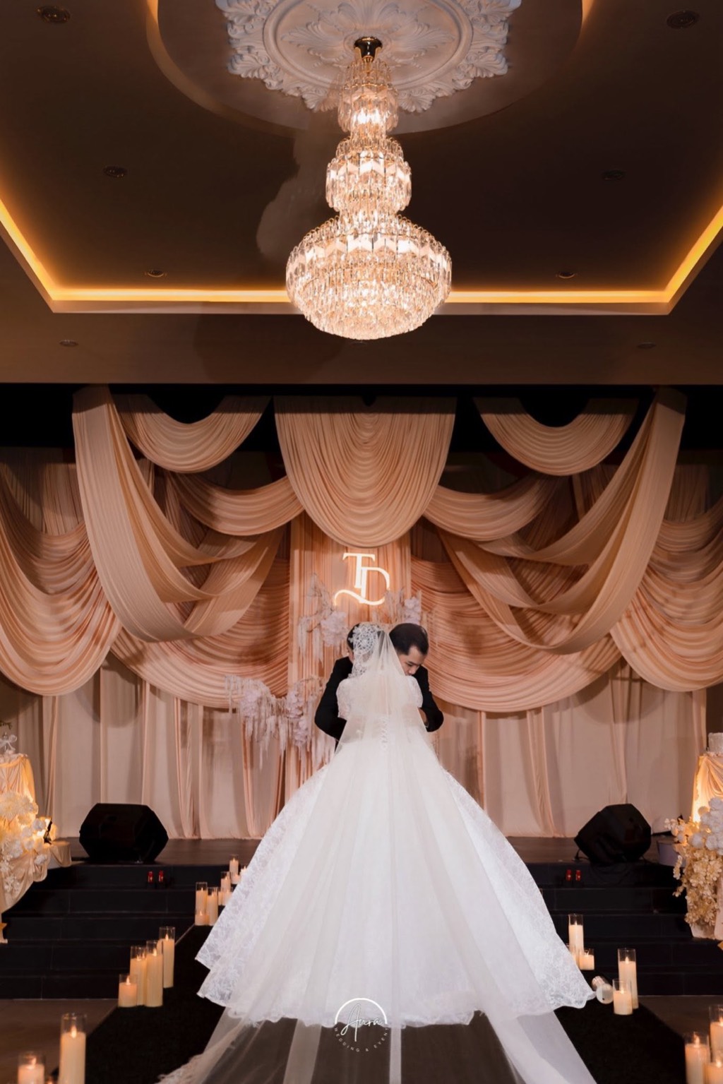 Cô dâu chú rể ôm nhau giữa lối nến, sân khấu rèm champagne và đèn chùm pha lê phía trên