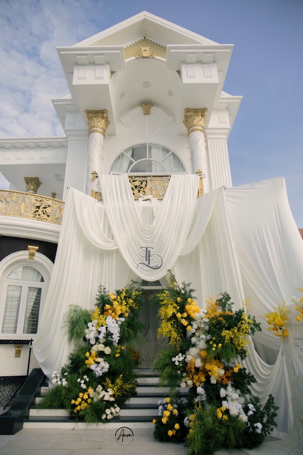 Mặt tiền biệt thự phủ lụa trắng rủ nhiều tầng với monogram TL, hai khối hoa vàng và trắng hai bên bậc thềm